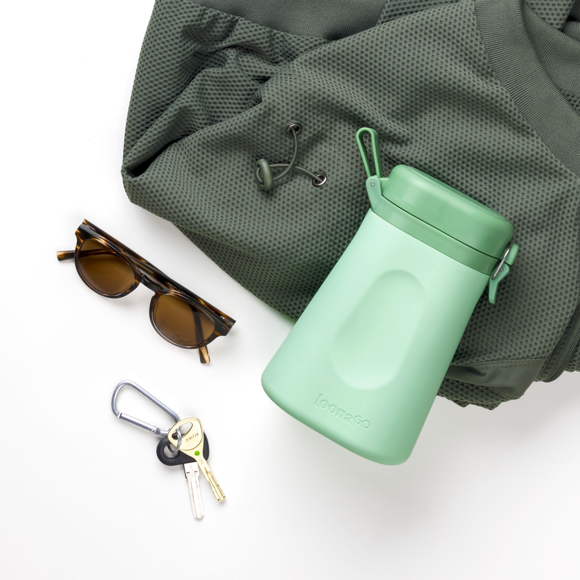 Mint green LoonaGo tucked into a green backpack alongside tortoiseshell sunglasses and keys, styled as an everyday carry essential