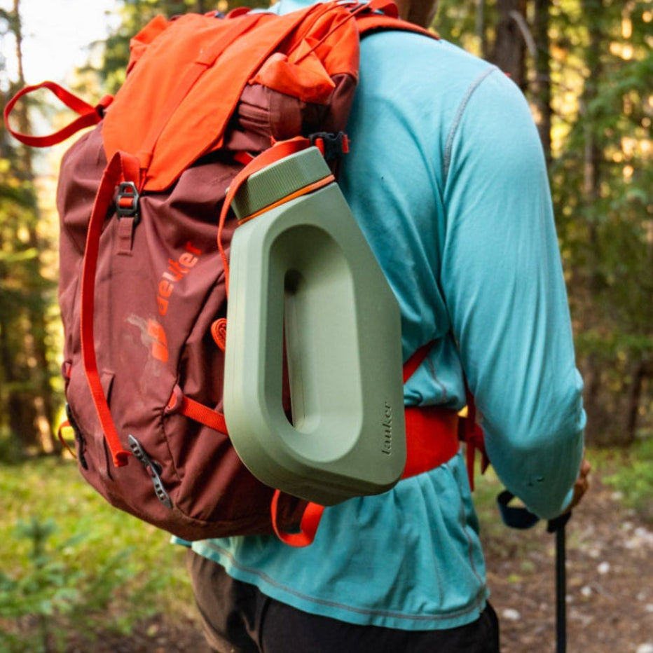 Sage green Tanker male urinal clipped to a hiker's red backpack on a forest trail, showing portability for outdoor adventures