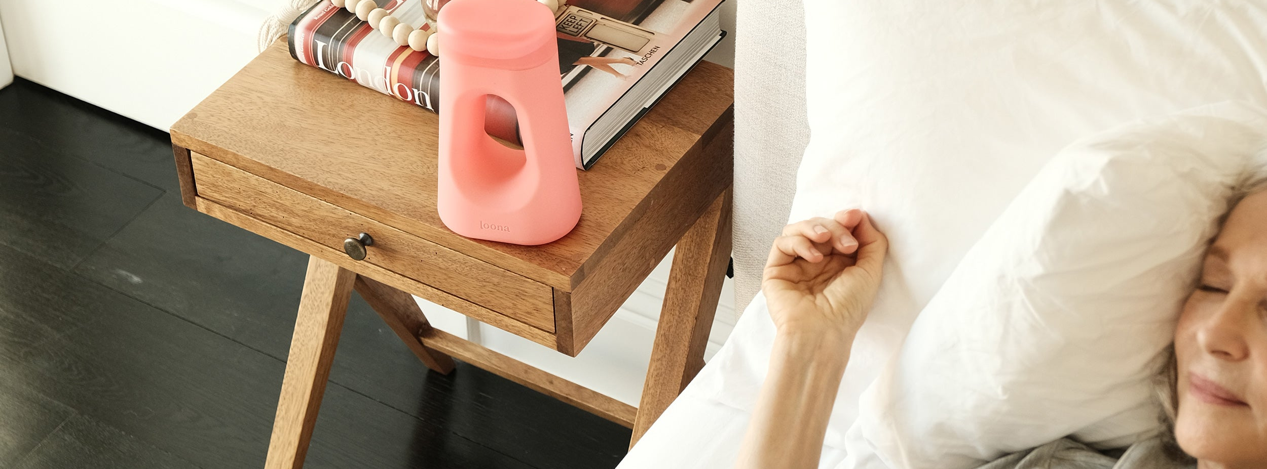 Coral Loona female urinal on a wooden bedside table with London art books, while a woman sleeps peacefully in bed nearby — illustrating its discreet overnight use