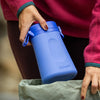 Woman in pink fleece dropping a blue LoonaGo female urinal into a green tote bag, showing its compact, portable size